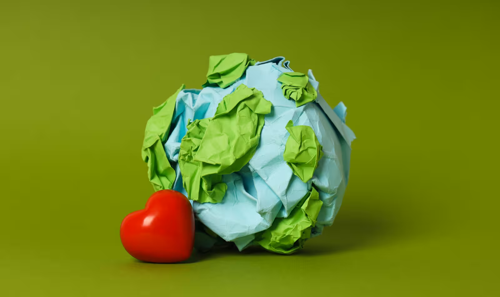 Globo terráqueo hecho con papel reciclado arrugado junto a un corazón rojo, sobre fondo verde